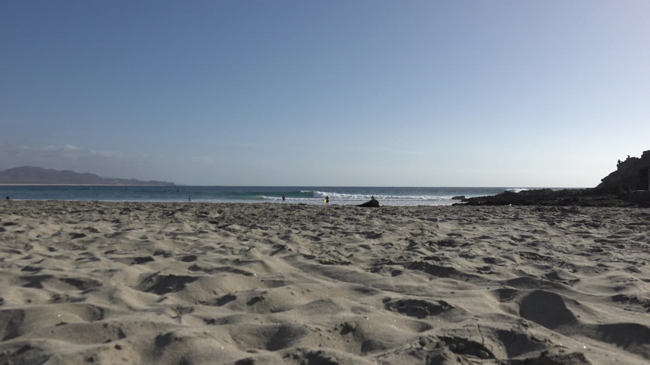 Los Cabos beach seen from low on rippled sand, with blue ocean waves, a rocky point on the right, hazy mountains on the horizon, and small distant people.