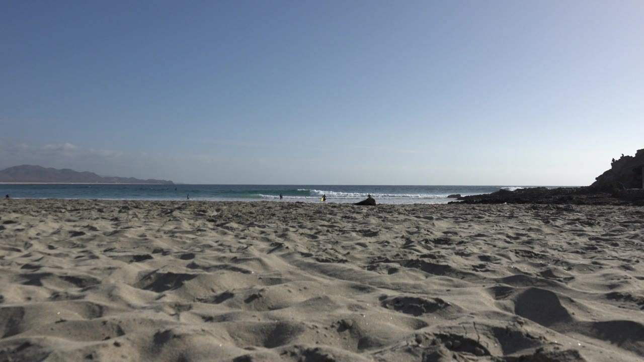 Los Cabos shoreline with textured sand in front, a band of breaking waves at mid-frame, distant mountains to the left, and tiny beachgoers near the waterline.