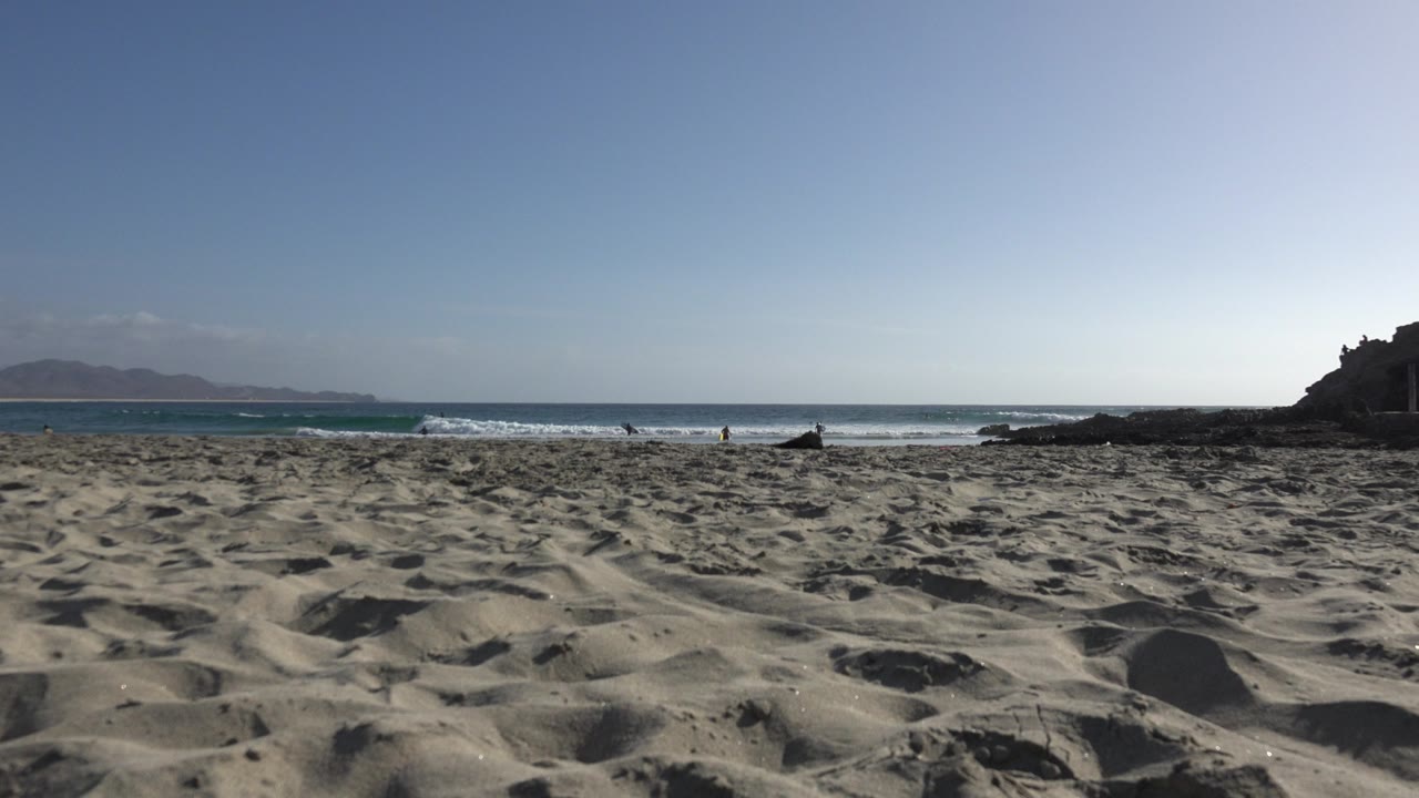 Los Cabos beach view one second in, with darker sand shadows in front, a line of white waves, distant land to the left, and the rocky shoreline holding the right side.