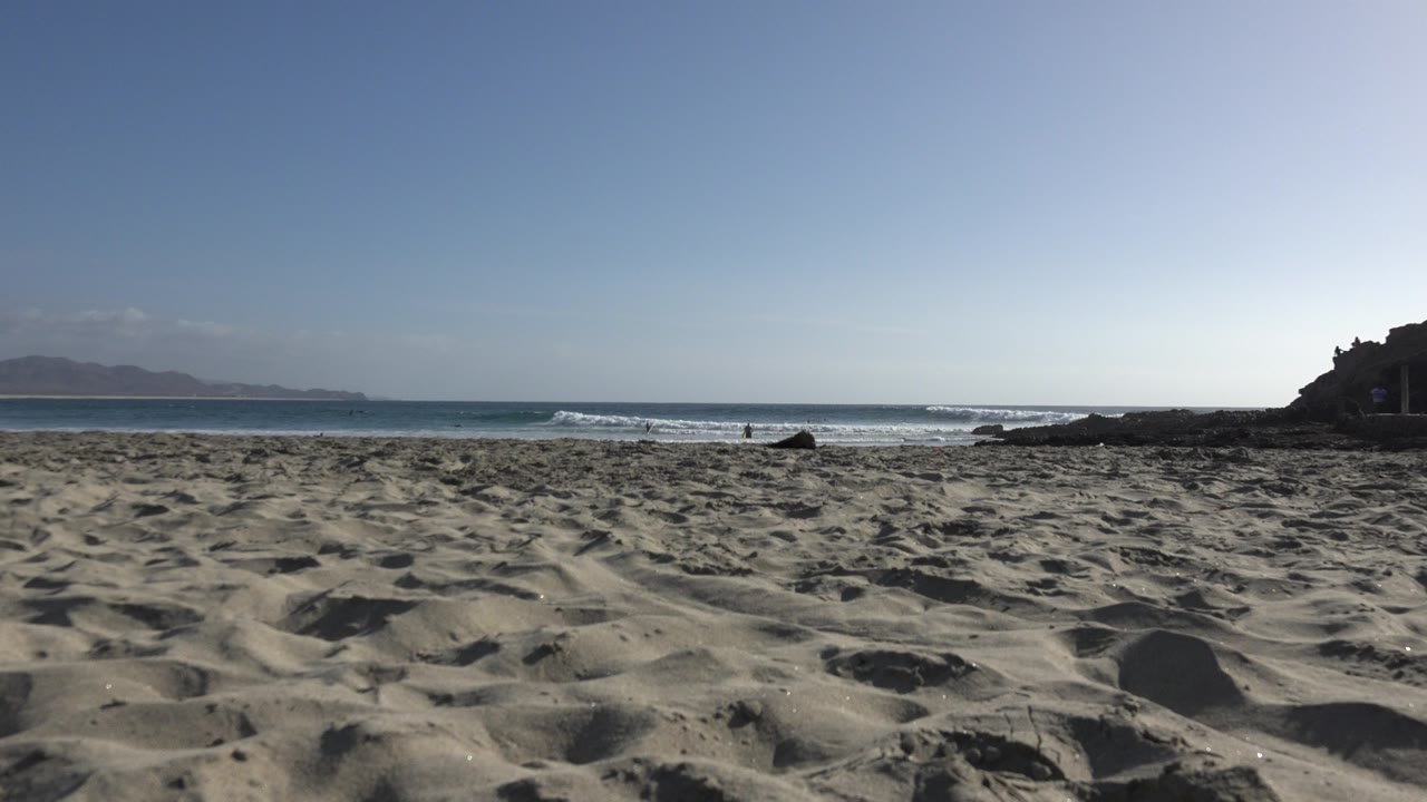 Low sand-level view of a Los Cabos beach with rippled foreground sand, breaking ocean waves, distant coastline on the left, dark rocks at the right edge, and no visible people.