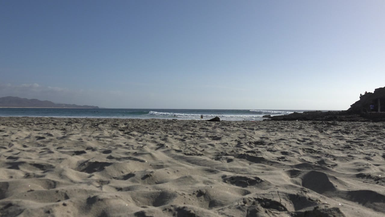 A second Los Cabos beach moment shows textured sand in front, a narrow band of blue water, small white surf, hazy distant land to the left, and no visible people.