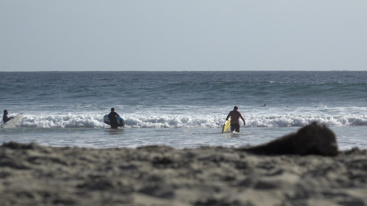 At 0:00 in Los Cabos, Mexico, distant beachgoers stand in shallow surf with boards while a low wave breaks behind them and blurred sand fills the foreground.