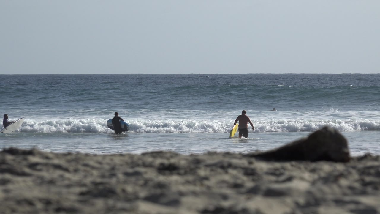 At 0:01 in Los Cabos, Mexico, several distant people are spread along the waterline as white surf pushes toward the beach under a pale sky.