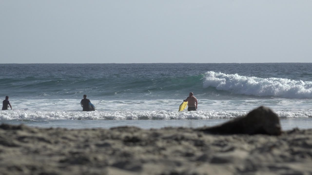 Los Cabos shoreline view with a blurred sandy foreground, breaking waves, and several distant people standing in shallow surf at the start of the clip.