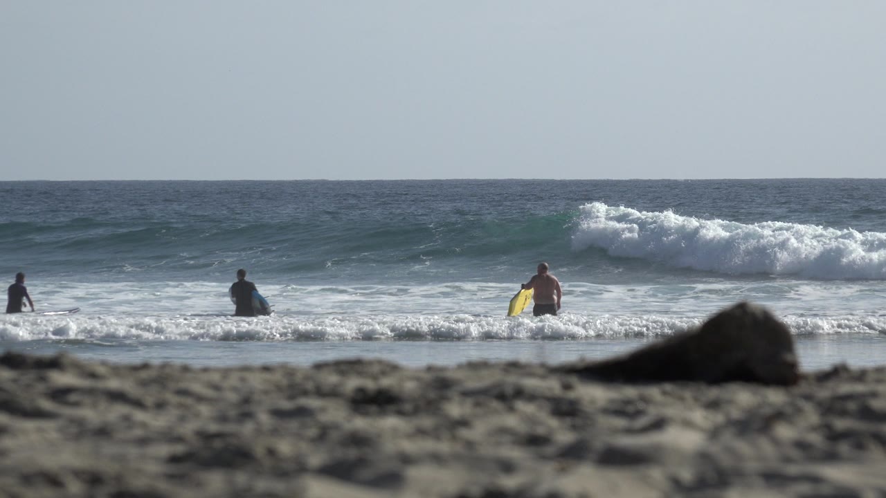 A white wave begins to break behind distant beachgoers in Los Cabos, with one person holding a bright yellow board near the center.