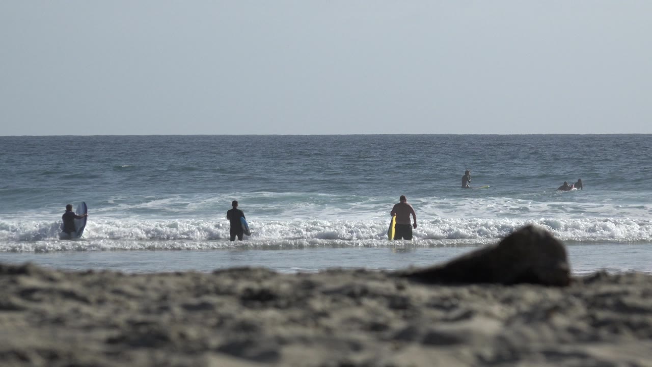 People standing and wading in surf behind blurred sand and a foreground rock.