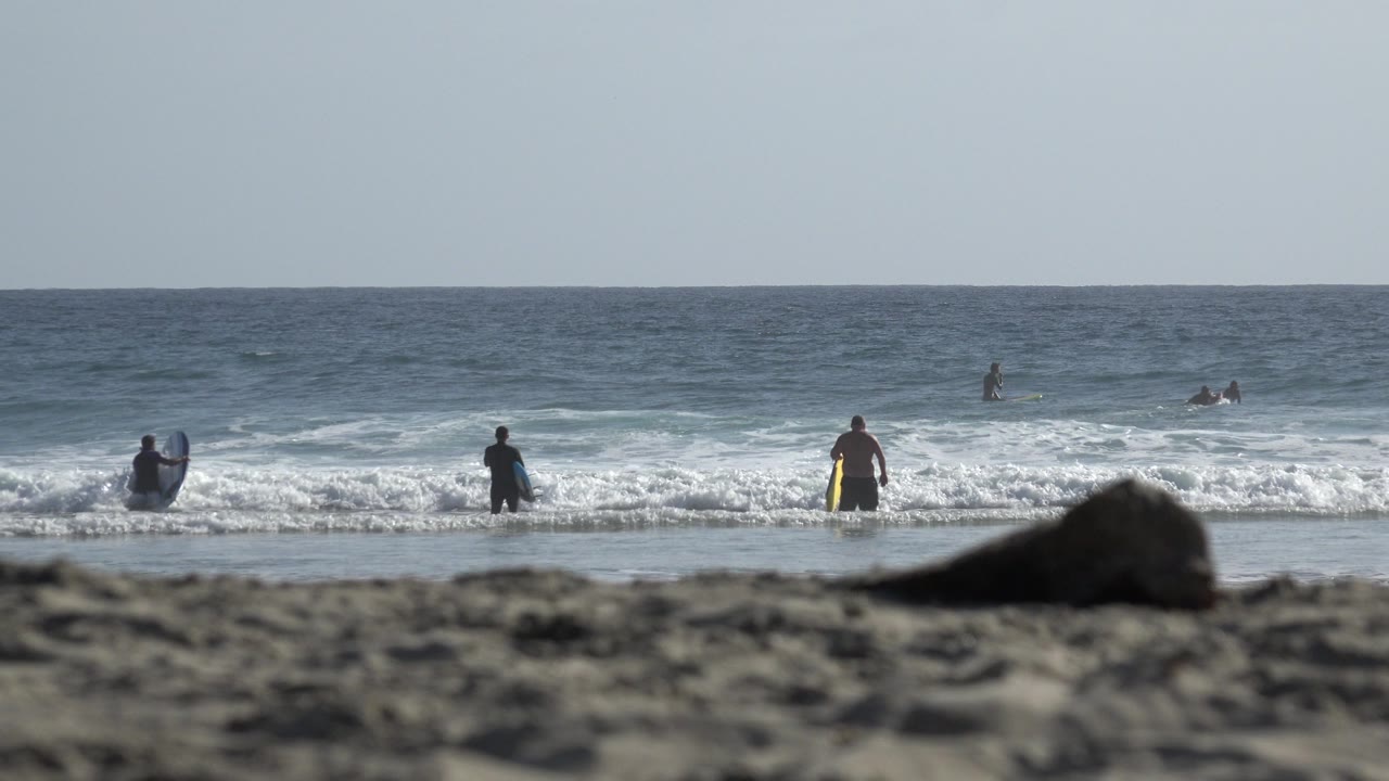 In Los Cabos, Mexico, small figures stand and wade along the white surf line while a person near center holds bright yellow swim fins or a board.