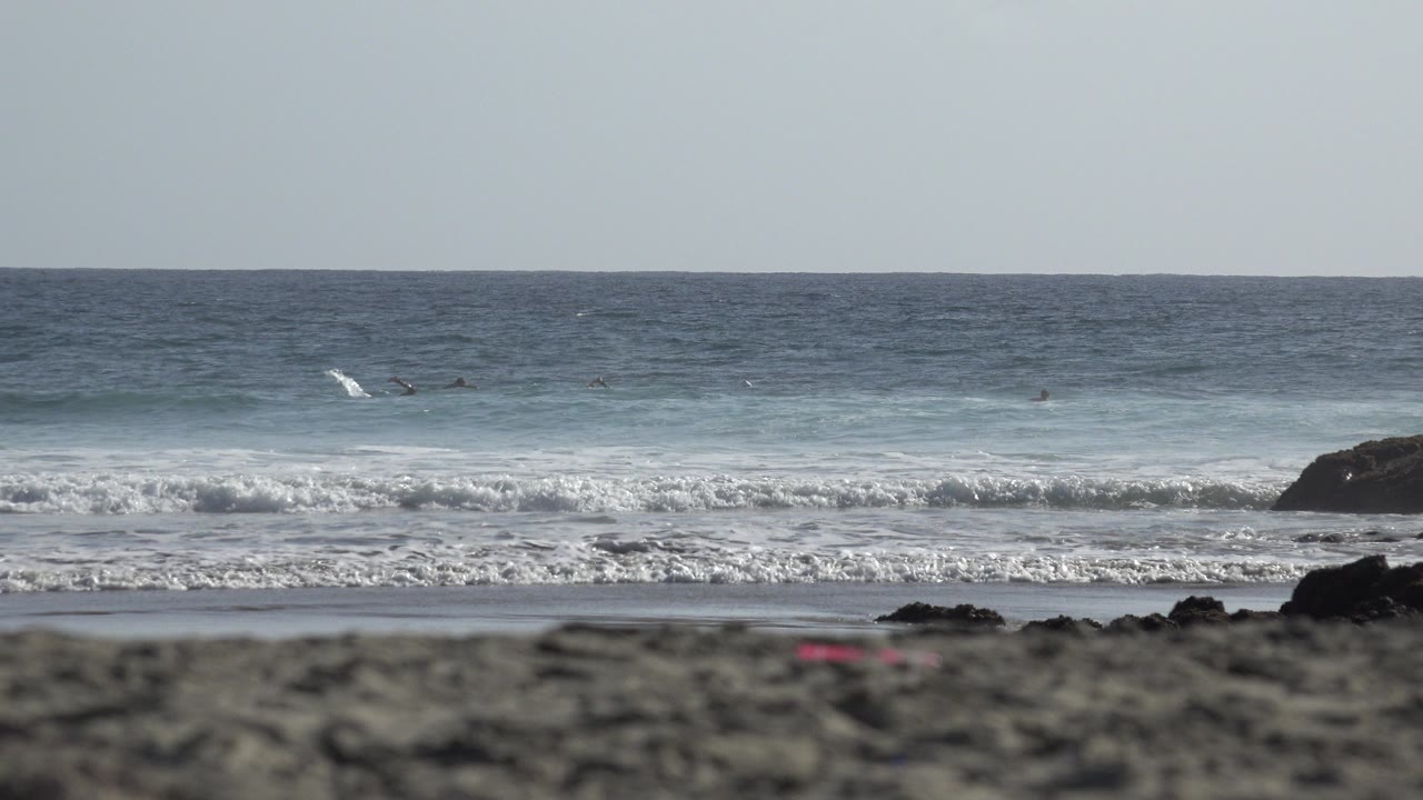 Los Cabos beach view with blurred sand in front, white surf rolling in, dark rocks near shore, and tiny surfers visible beyond the first break.
