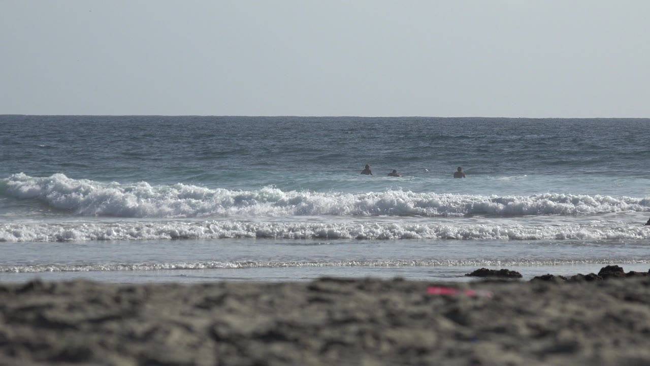 A low Los Cabos shoreline frame shows foamy waves advancing toward wet sand while small distant figures remain scattered beyond the break.