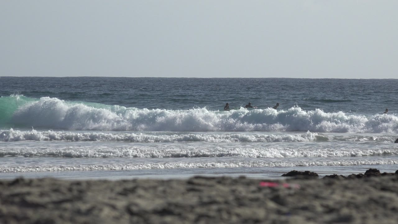Los Cabos surf scene with a higher white wave crest in the midground, dark blurred beach foreground, flat ocean horizon, and small distant figures partly hidden by spray.