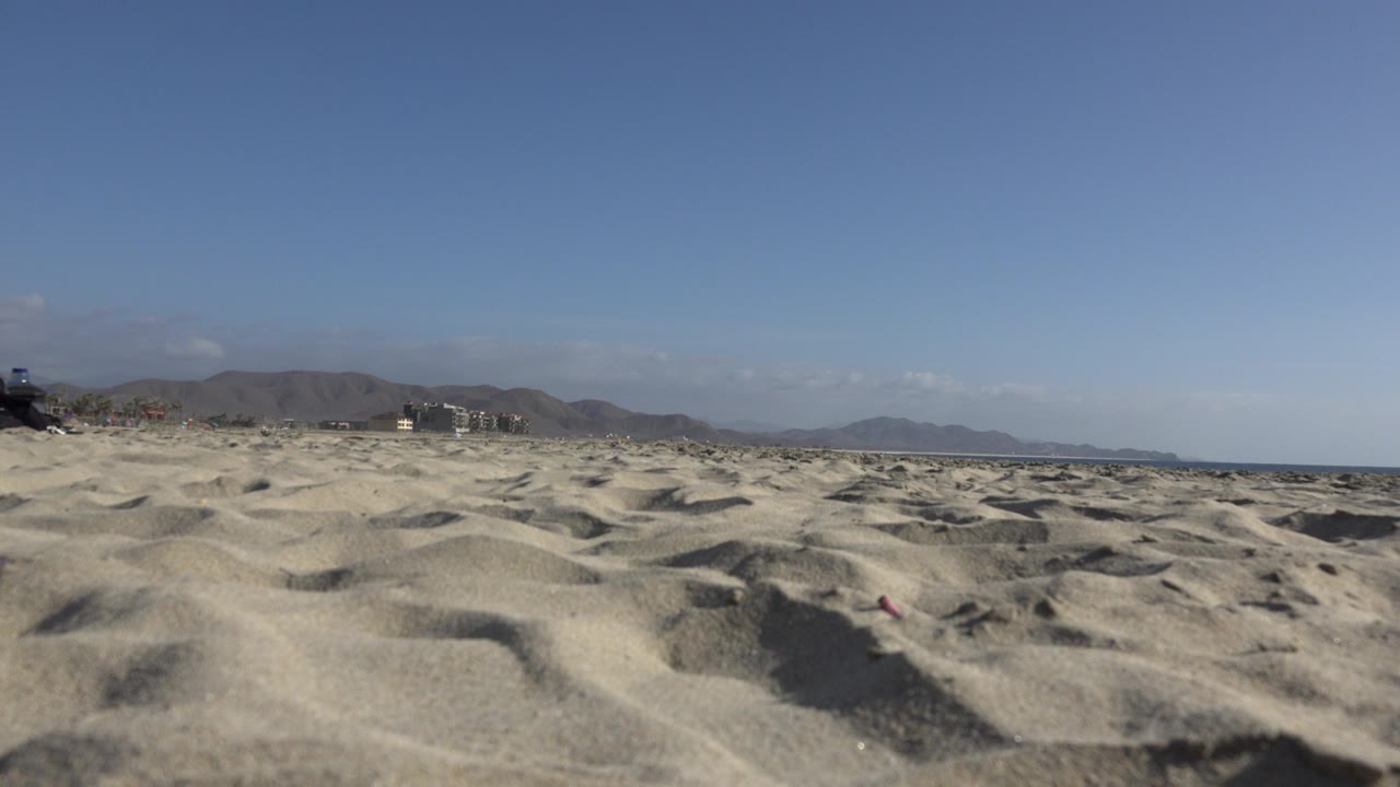 Low foreground sand ridges stretch toward distant hills and small buildings in Los Cabos, Mexico, with blue sky above and no visible people.