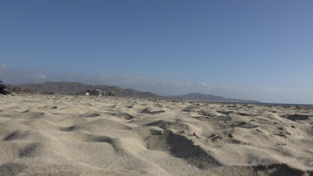 Rippled beach sand with sea, distant buildings, mountains, and blue sky.