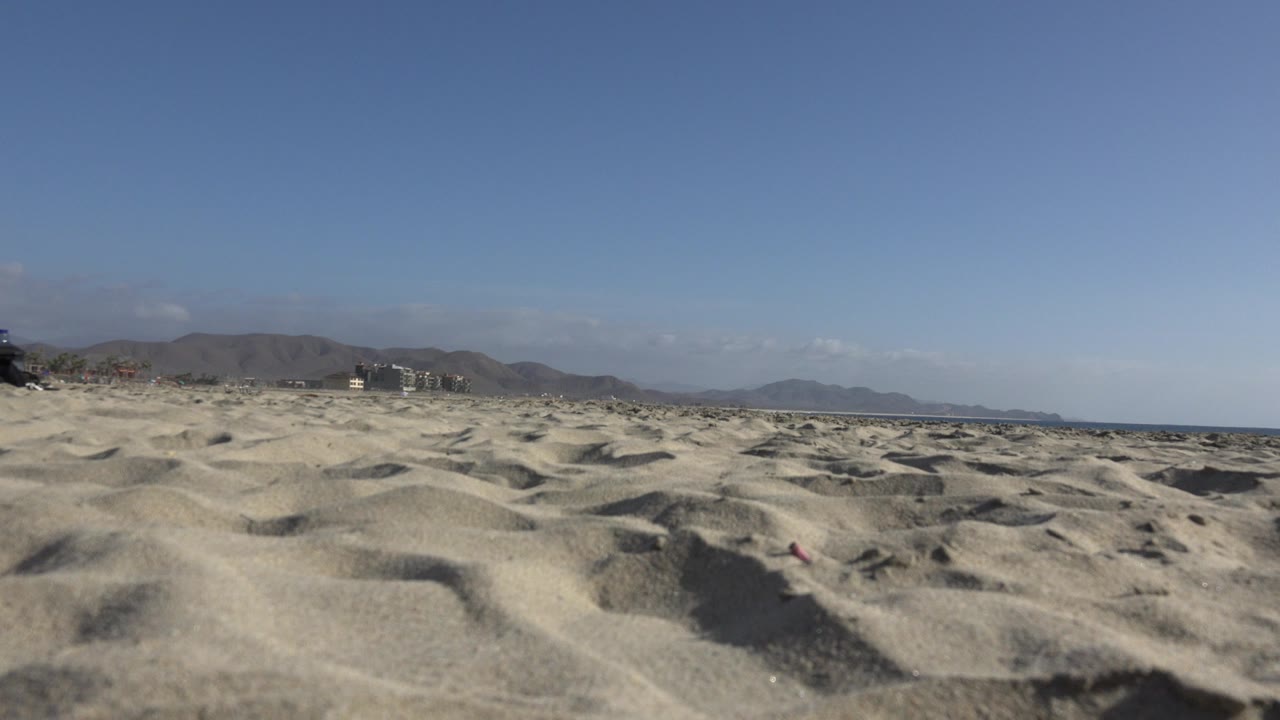 Low-angle view of rippled sand dunes in Los Cabos, Mexico with distant brown mountains, small horizon buildings, blue sky, and no visible people.