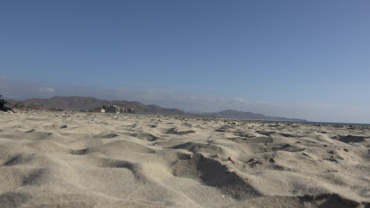 Opening Los Cabos beach frame with rippled sand in the foreground, distant mountains along the horizon, a narrow ocean line, and no visible people.
