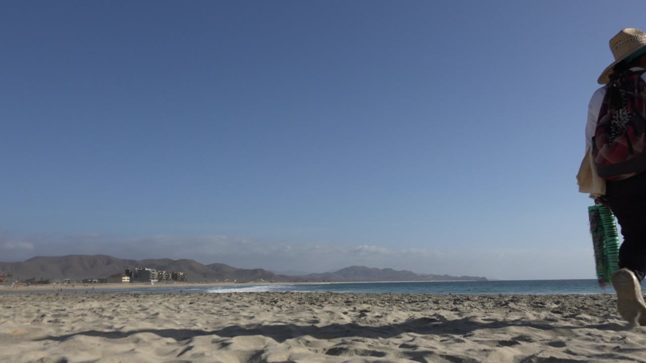 A person in a wide hat and dark backpack walks farther into the Los Cabos beach frame, with rippled sand in front and a low mountain line beyond the water.