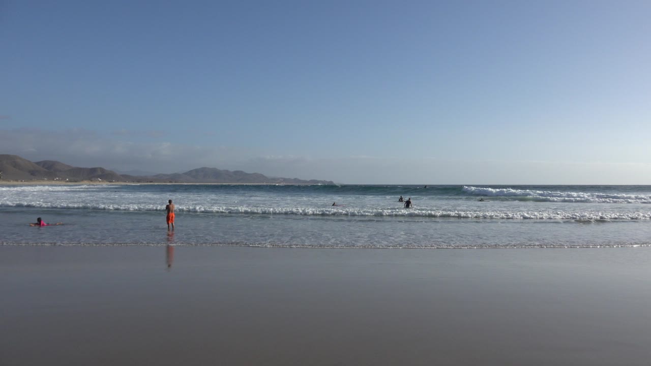 Wide beach view with waves, wet sand, distant hills, and a few people near the water.