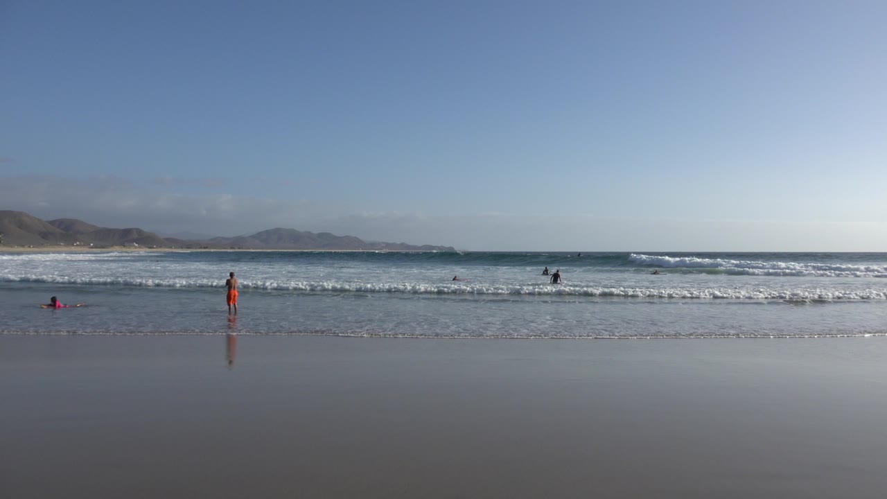 At 0:01, the Los Cabos shoreline remains wide and open as small figures stand in the water while a pink board or float sits near the left surf line.