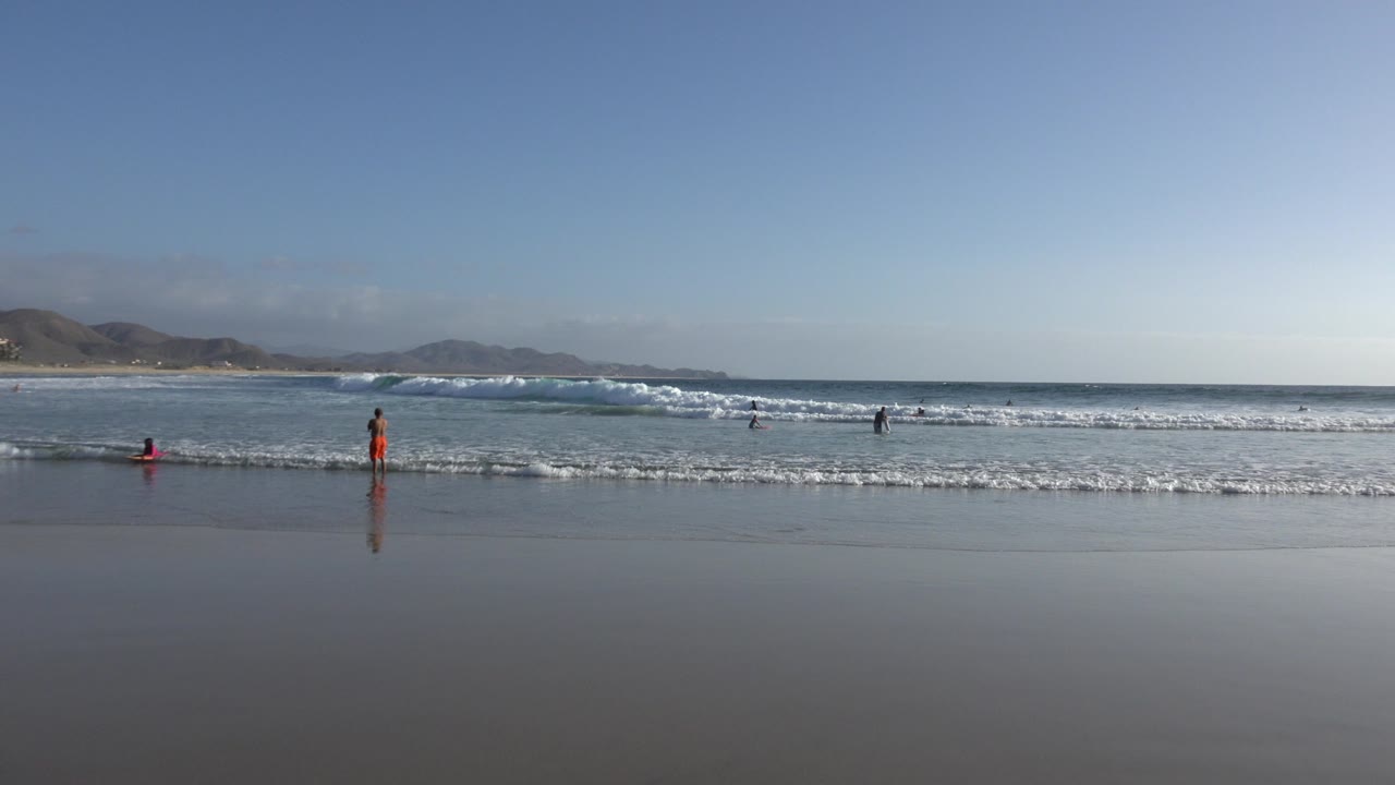 Wide Los Cabos beach view with wet reflective sand, small people in the shallow surf, white waves, pale blue sky, and distant mountain silhouettes.