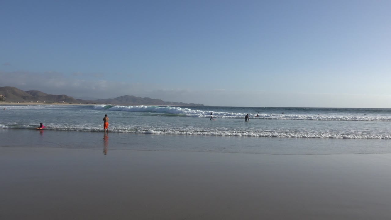 Los Cabos shoreline frame with a person in orange near the center, other beachgoers spread across the surf, white foam bands, and mountains in the distance.