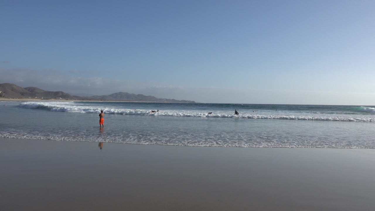 Wide beach view with surf, wet sand, distant hills, and small figures near the water.