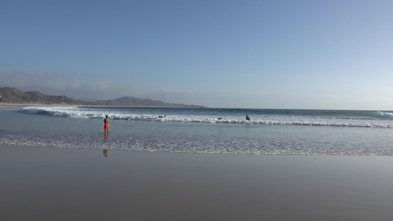 A second view of the Los Cabos shoreline shows the surf line slightly shifted, with wet sand reflecting the sky and small distant figures spaced along the water.