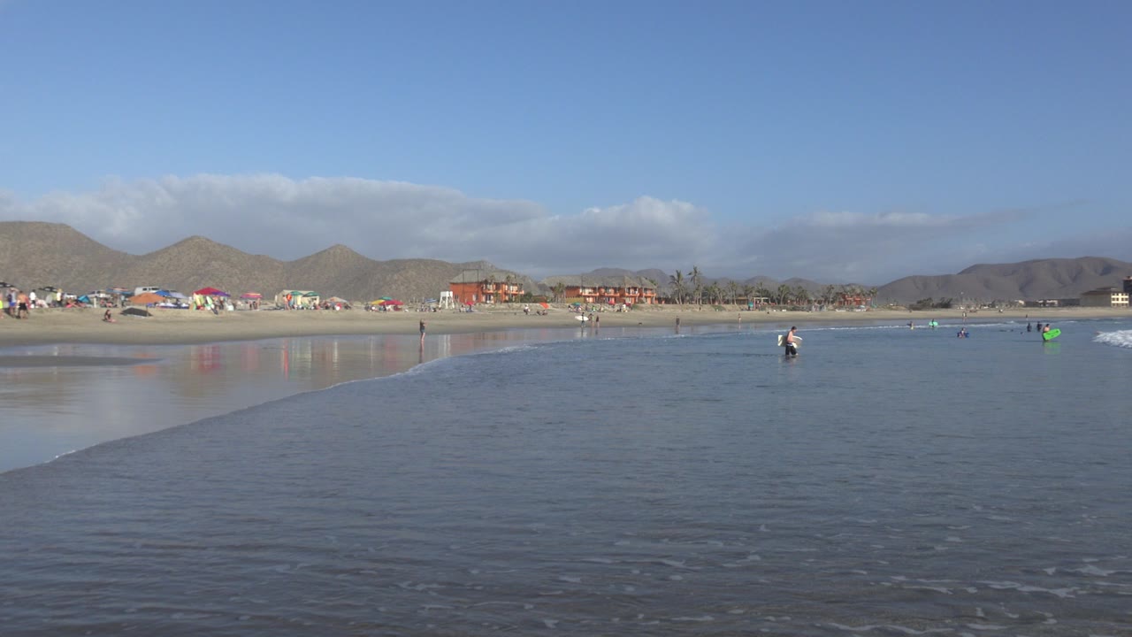 Wide Los Cabos beach frame with rippled shallow water in the foreground, small beachgoers along the shore, low buildings, brown hills, and a bright blue sky.