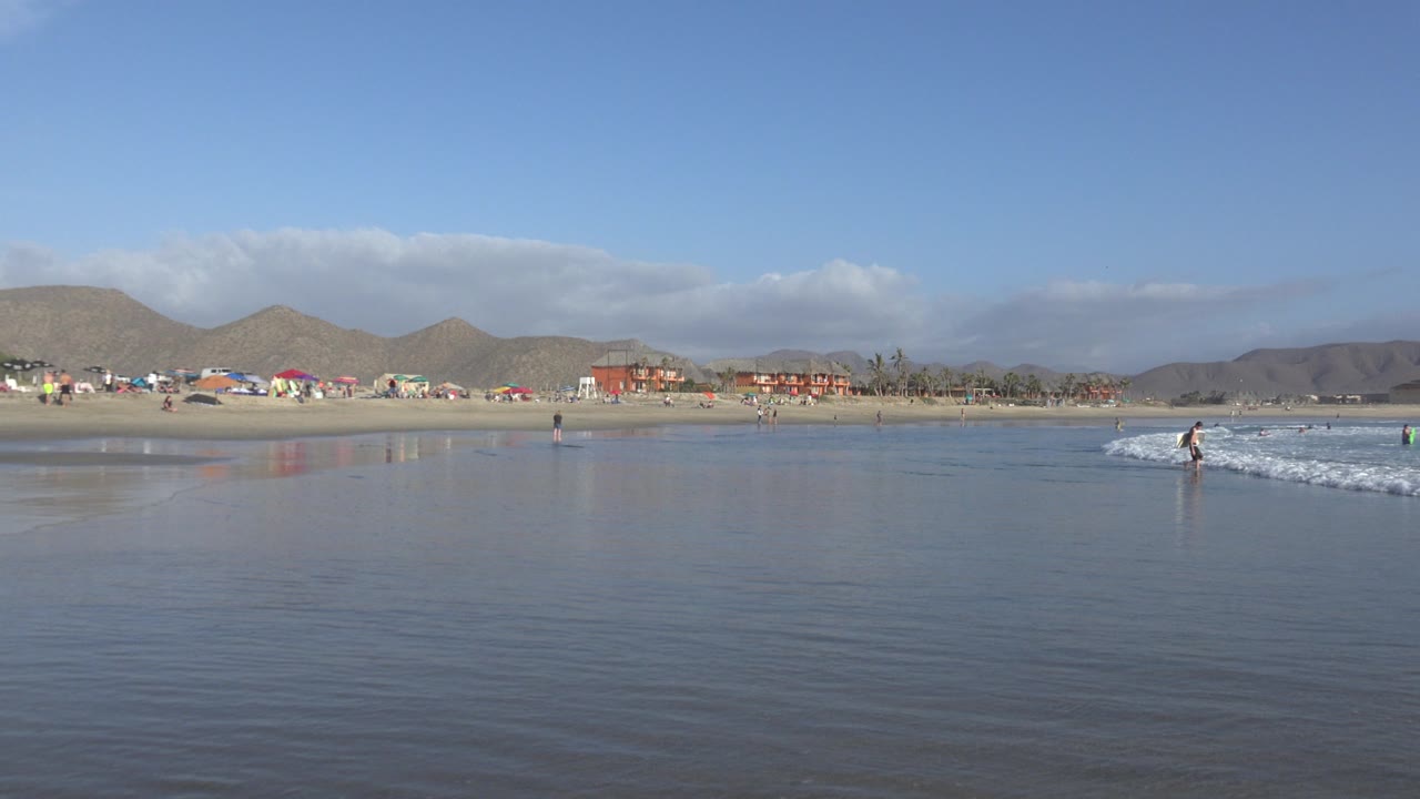 Wide view of shallow water at a Los Cabos beach, with distant umbrellas, small beachgoers, low hills, and bright blue sky.