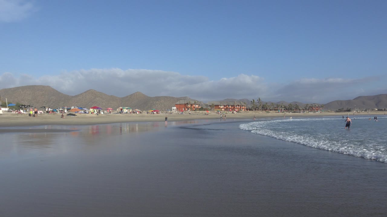 Wide Los Cabos beach view with reflective wet sand in front, a low wave entering from the right, umbrellas and low buildings in the distance, and small beachgoers near the shoreline.