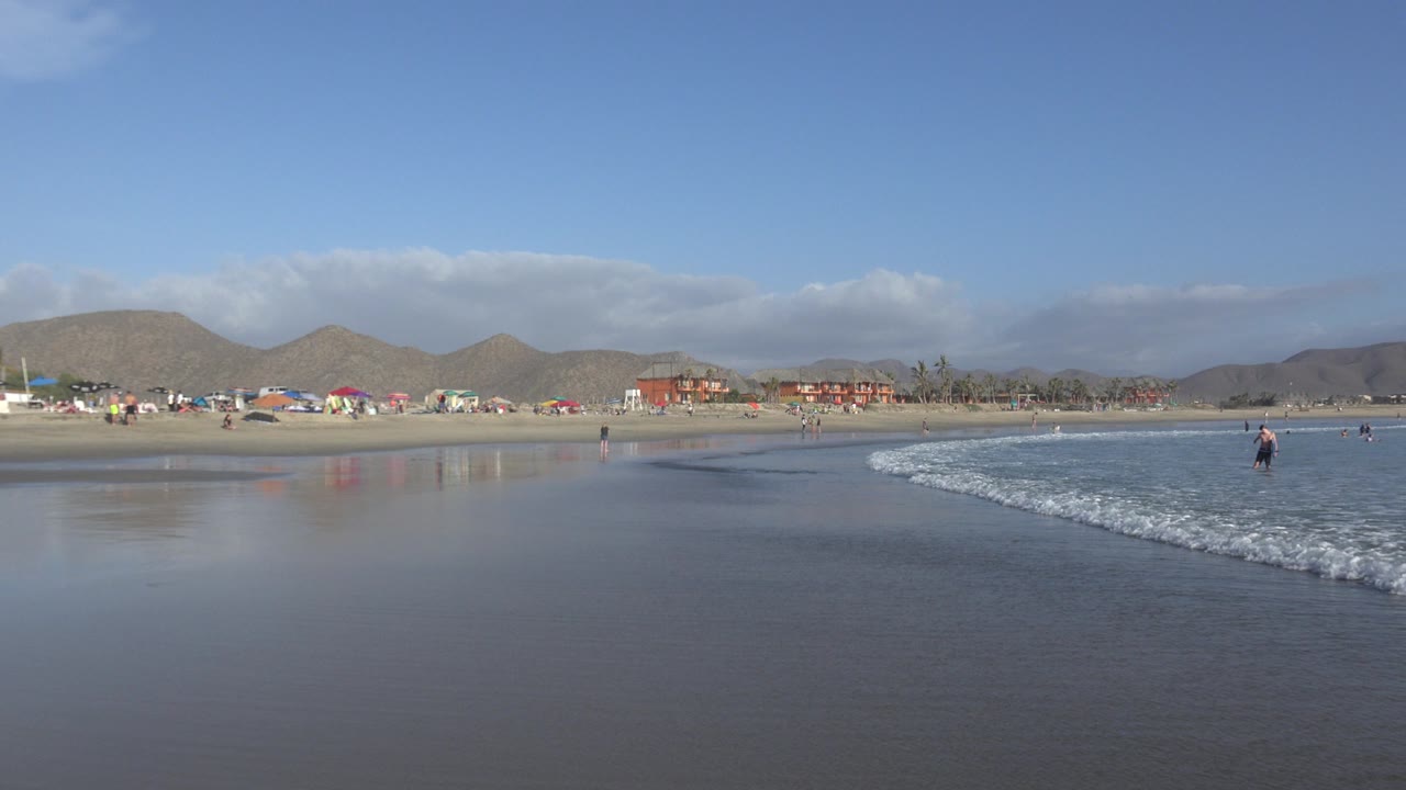 Los Cabos shoreline with the white foam line farther across the wet sand, distant umbrellas and buildings along the beach, hills behind them, and small figures near the water.