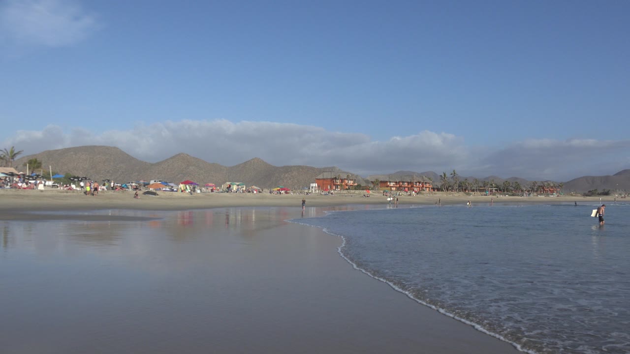 Los Cabos shoreline with a small person standing near the waterline, reflective sand in the foreground, beach umbrellas and low buildings below brown hills.