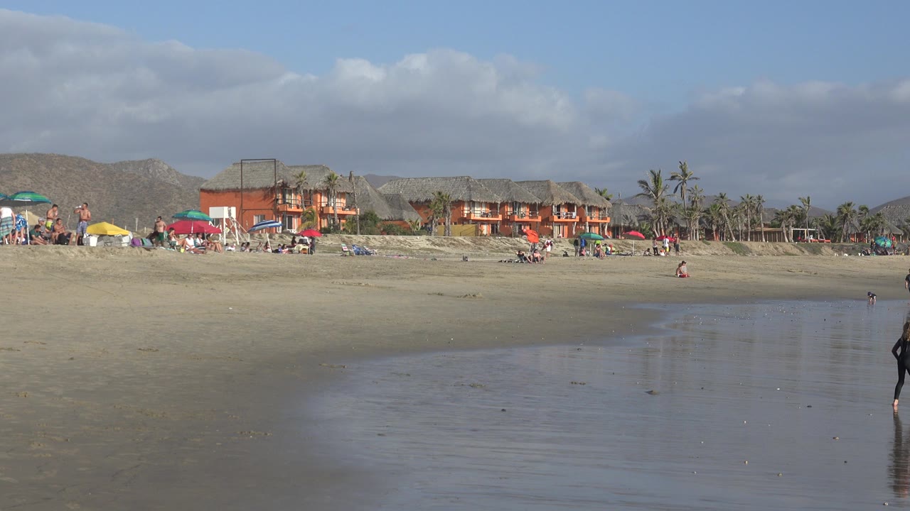 Wide cloudy beach with wet sand, distant umbrellas, buildings, palms, and scattered people.