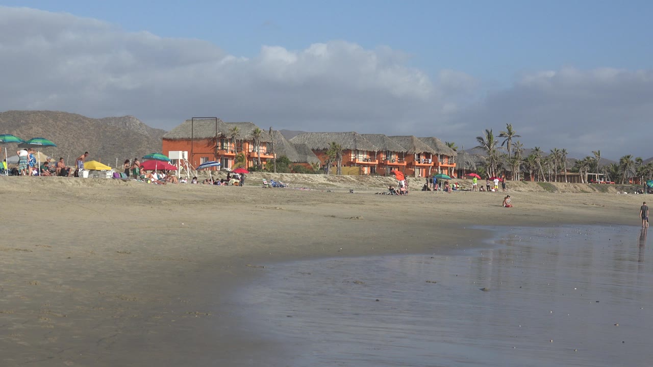 Wide beach with shallow water, umbrellas, people, buildings, palms, and hills.