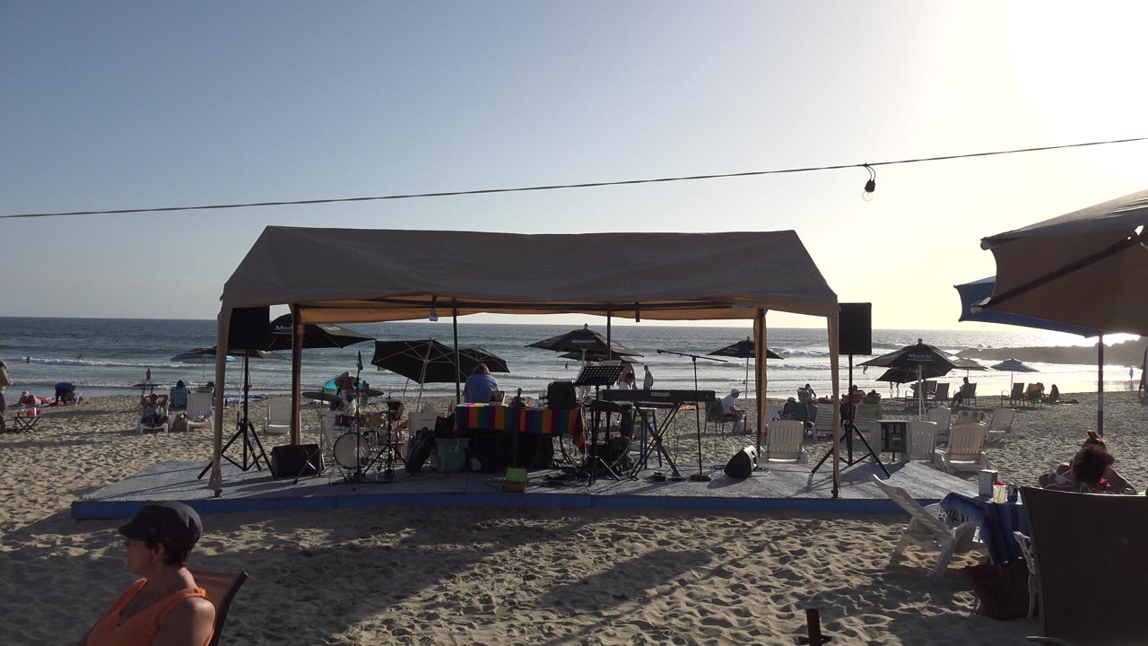 Beach canopy with equipment, umbrellas, seated people, and ocean waves.