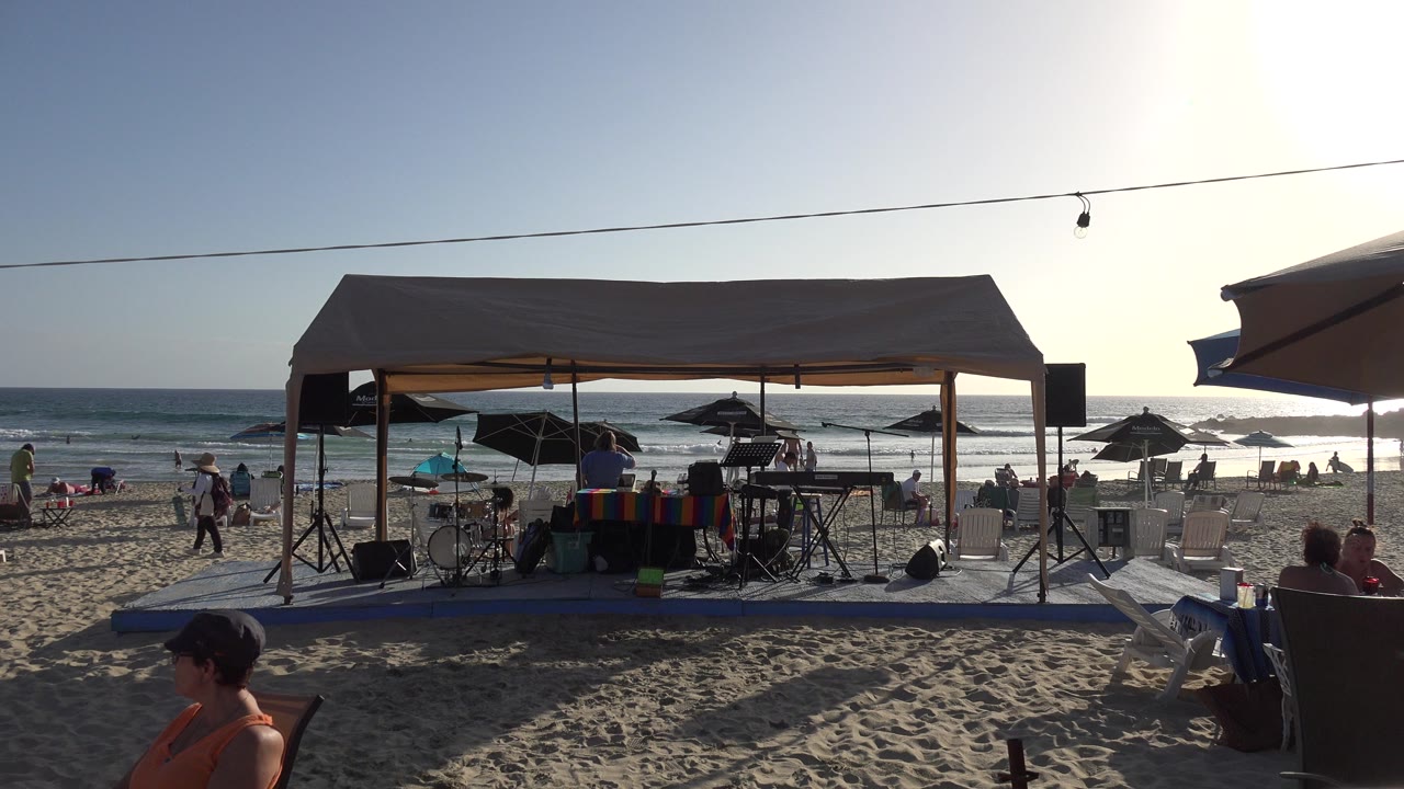 Beach canopy with music equipment, speakers, umbrellas, ocean waves, and people on sand.