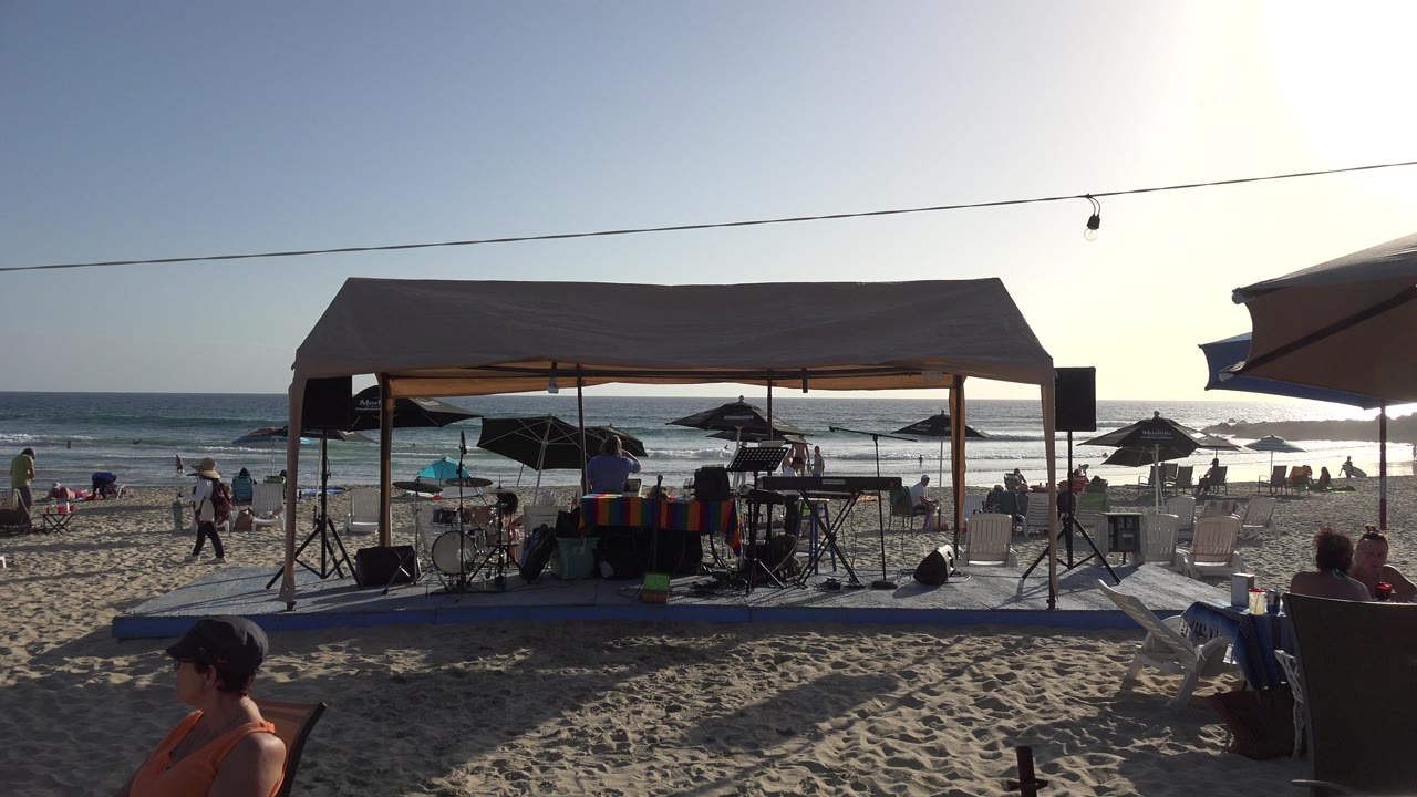 At 0:01, the Los Cabos beach canopy remains centered with music equipment under shade, while people sit and walk near umbrellas by the waterline.