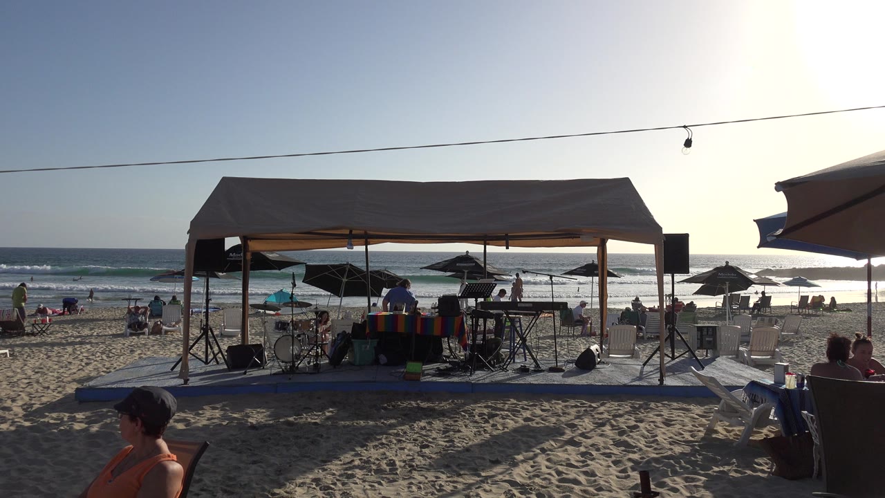 At 0:01 in Los Cabos, the canopy and audio gear remain centered while beachgoers sit and move near umbrellas with ocean surf in the background.