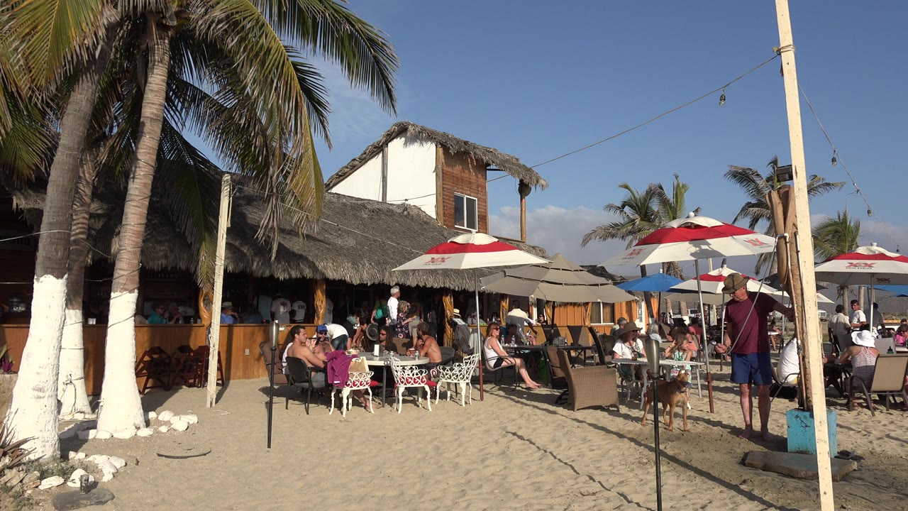 Beach restaurant with umbrellas, palm trees, and diners on sand.