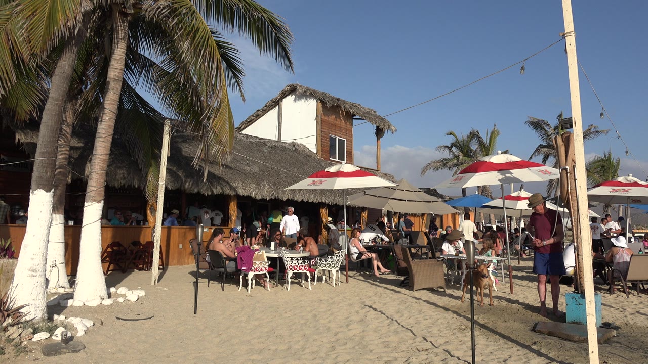 Opening frame of a Los Cabos beach seating area with people at tables on sand, palm trunks at left, red-and-white umbrellas, and a thatched structure behind them.