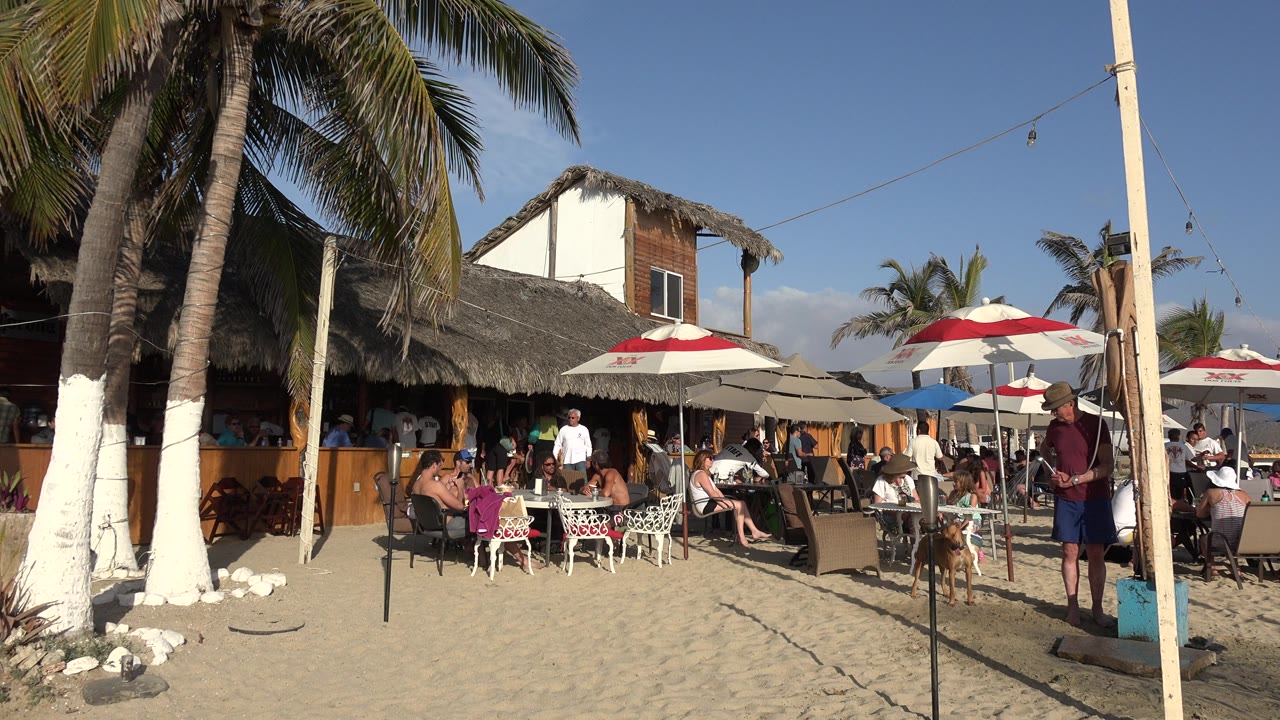 Los Cabos beach dining area one second in, with the same seated group visible under shade umbrellas and a standing man near a pole on the right edge.