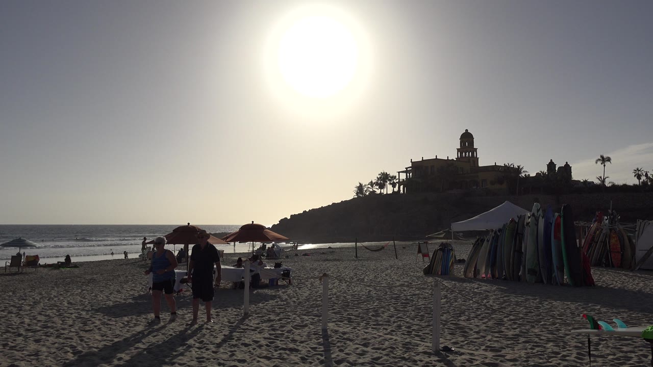 Backlit beach with tourists, umbrellas, surf gear, ocean, and hilltop building.