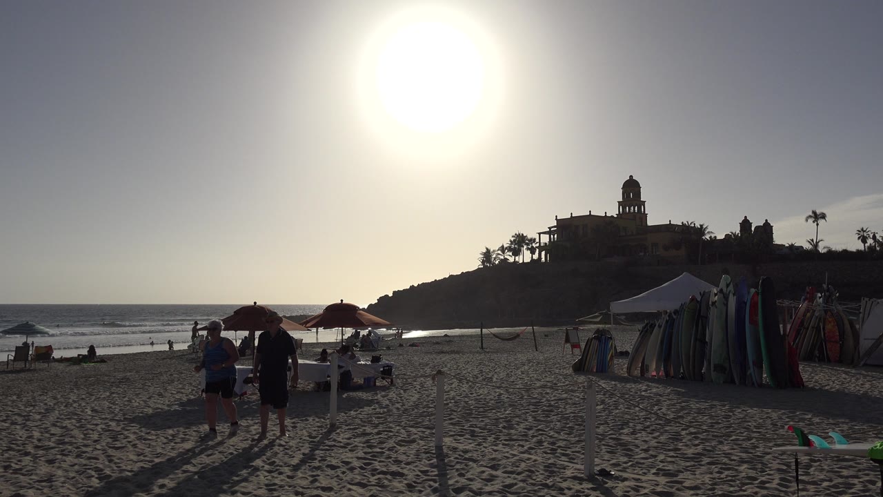 Los Cabos beach scene with tourists moving near the center of the frame, surfboards or beach gear lined beside a tent, umbrellas by the shoreline, and ocean waves in backlight.