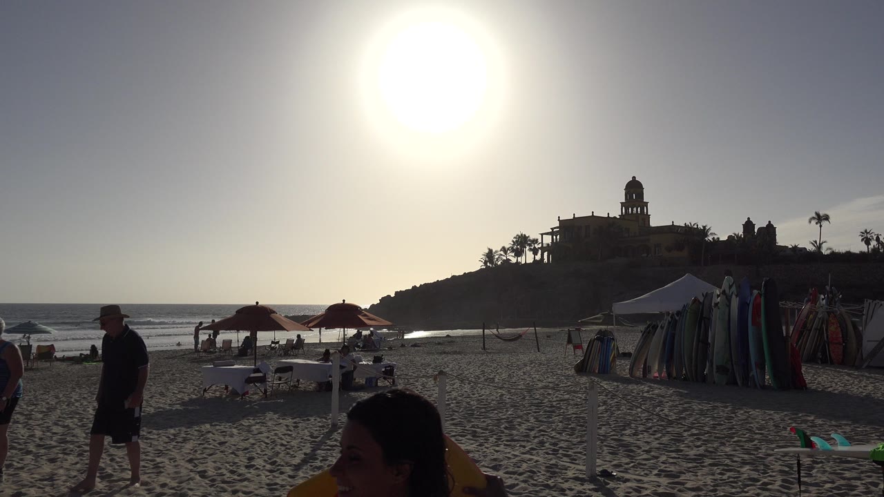 Los Cabos beach view at the start of the clip, with bright sun overhead, umbrellas on the sand, surfboards near a canopy, ocean waves, hillside buildings, and visible people in the foreground and midground.