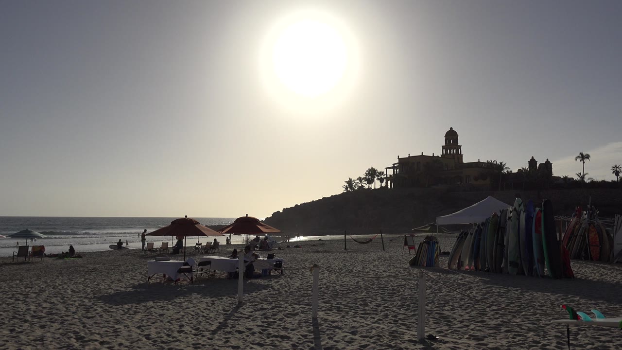 Los Cabos shoreline one second in, showing a high white sun over beach umbrellas, clustered tents, textured sand, waves, and small beachgoers in silhouette.