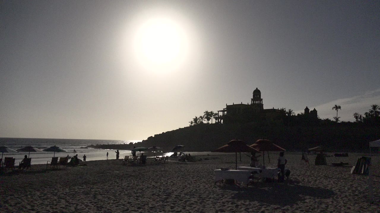 Backlit beach with bright sun, umbrellas, ocean, and silhouetted hill.