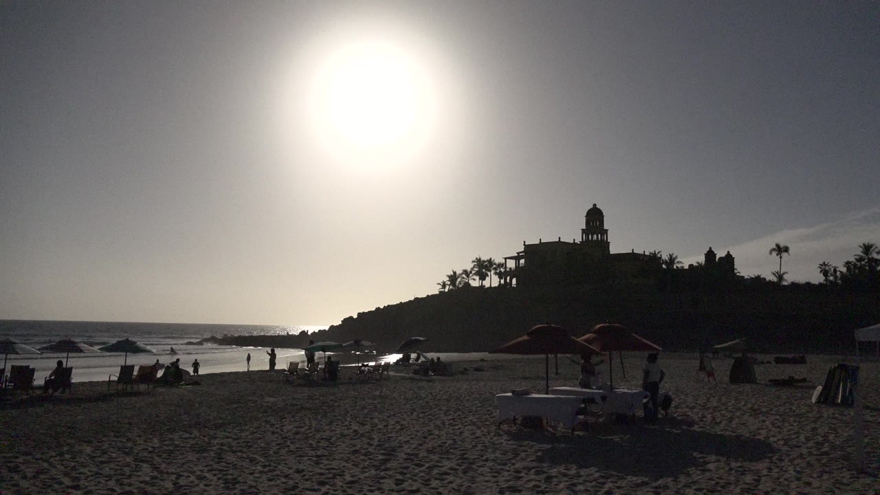 Los Cabos beach one second in, with intense sun flare above the sand, small bathers near the waterline, shaded umbrellas in the midground, and the dark hillside building still prominent.