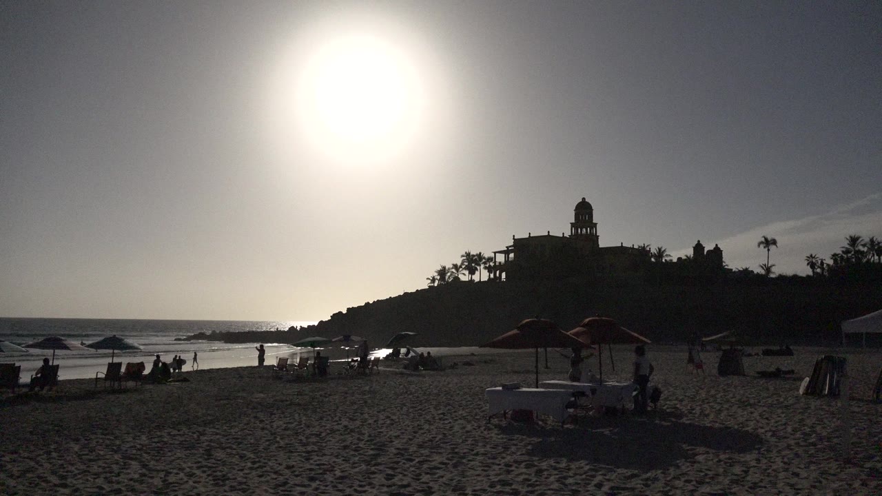 Backlit beach with umbrellas, people, ocean, and a hilltop building silhouette.