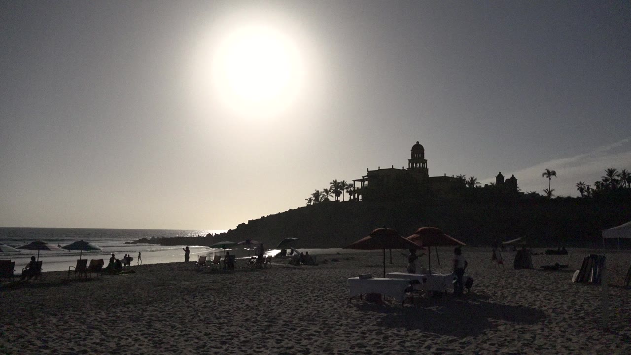 Los Cabos beach frame at 0:01 with sun glare above the shoreline, dark beach furniture in the foreground, people near the waterline, and the hilltop building in silhouette.