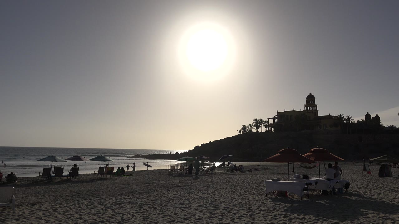 Backlit beach umbrellas, lounge chairs, ocean, and a hilltop building silhouette.