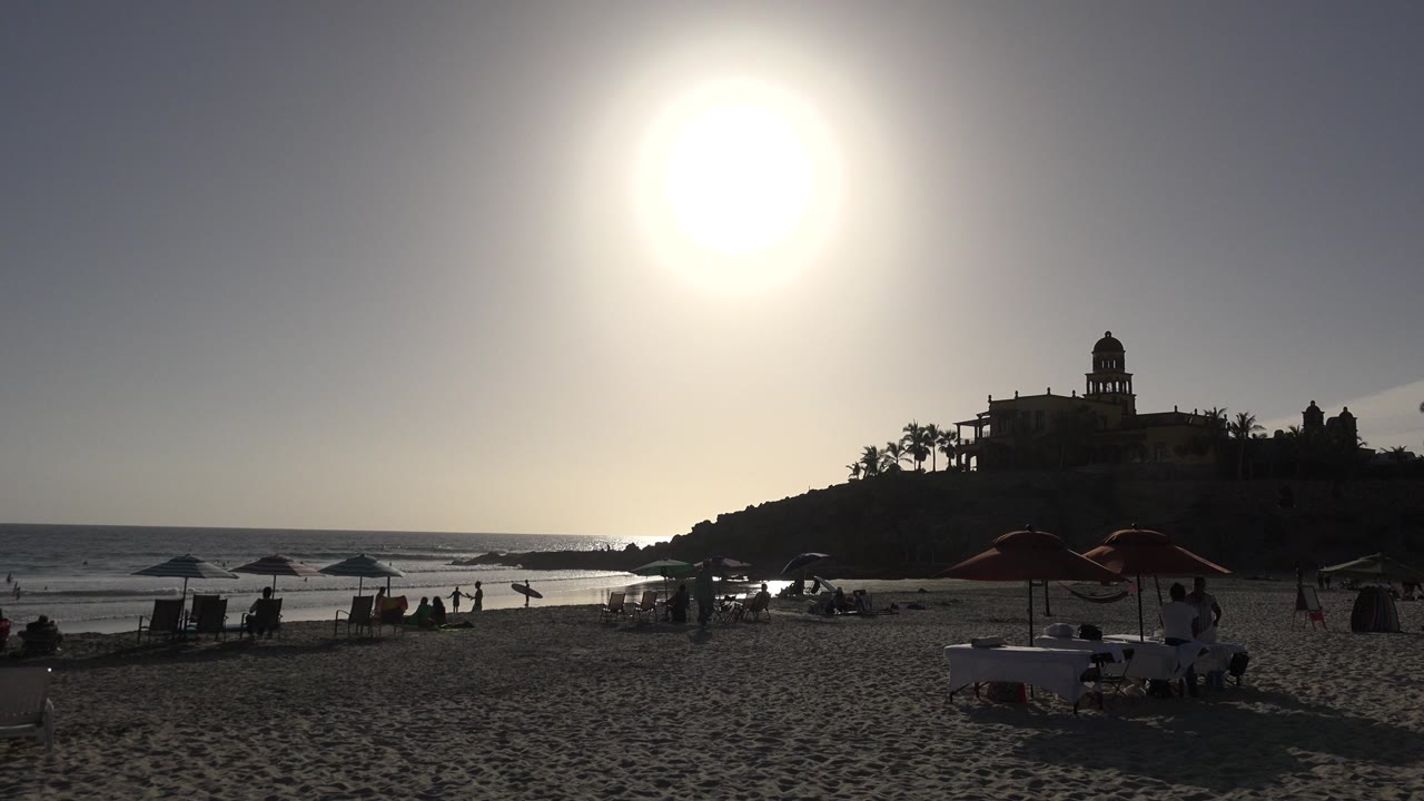 Los Cabos shoreline scene with beach chairs and umbrella shade in the foreground, several small beachgoers near the water, and a silhouetted building on the rocky point.