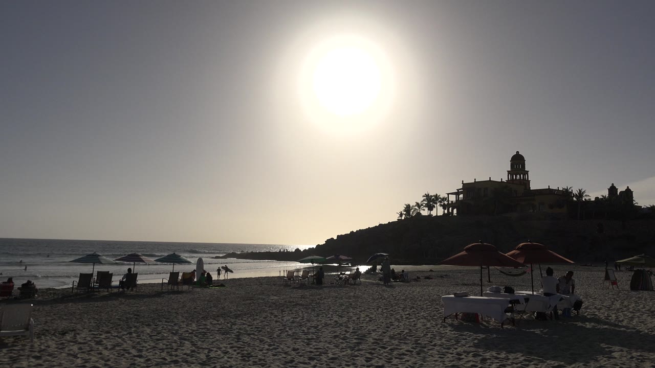 Bright sun over a sandy beach, ocean, parasols, and silhouetted hilltop buildings.